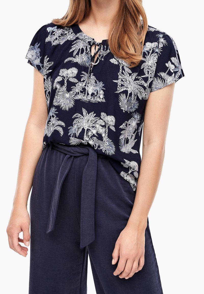 Femme portant un pantalon bleu marine avec ceinture en tissu et un chemisier bleu marine à manches courtes avec des imprimés blancs de plantes tropicales et un nœud au niveau du col.
