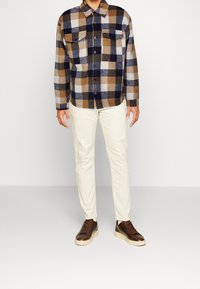 En flanellskjorta i brun, marinblå och krämfärgad rutmönster med två bröstfickor, kombinerad med vita byxor och mörkbruna sneakers.