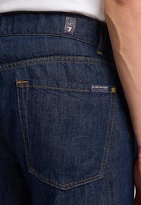 Jeans in denim blu scuro con cuciture arancioni, dotati di un'etichetta sulla tasca posteriore e di un'etichetta visibile in vita impressa con il numero 7.
