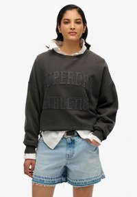 Sweatshirt gris foncé avec "SUPERDRY ATHLETIC" en lettres en relief, porté sur une chemise blanche à col, associé avec un short en denim bleu clair.