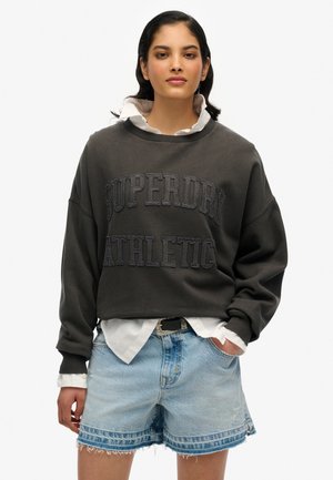 Sweatshirt gris foncé avec "SUPERDRY ATHLETIC" en lettres en relief, porté sur une chemise blanche à col, associé avec un short en denim bleu clair.