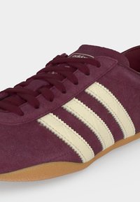 Adidas sneaker από σουέντ σε μαρόν χρώμα με κρεμ τρεις ρίγες, μαρόν κορδόνια, και σόλα από καουτσούκ, με κοντινή λήψη πλευρικών και κορυφαίων λεπτομερειών.