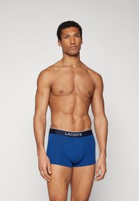 Kék pamut boxer alsónadrág fekete derékkal, amelyen fehér "LACOSTE" felirat látható. Szoros illeszkedésű kialakítás, sima tapintású.