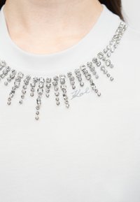 Λευκό τοπ με ψηλό λαιμό και καταρράκτη διαφανών στρας γύρω από το ντεκολτέ, με μια διακριτική κεντημένη υπογραφή στο στήθος.