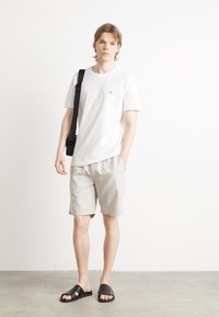 Tricou alb cu mânecă scurtă și un logo bleumarin, pantaloni scurți bej deschis și șlapi negri. Țesătură netedă cu o croială lejeră.