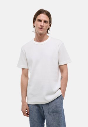 Jeune homme aux cheveux bruns mi-longs portant une chemise blanche texturée à manches courtes et un pantalon bleu sur un fond uni.