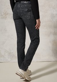 Jeans de mezclilla negros con corte ajustado, con bolsillos traseros y una textura deslavada. Combinados con zapatillas deportivas de color claro sobre un fondo neutro.