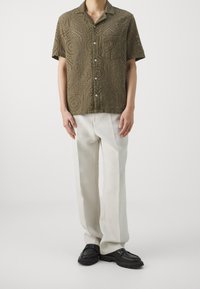 Korte mouwen, olijfgroen overhemd met een gestructureerd, uitgesneden patroon en een borstzak, gecombineerd met lichtbeige broeken en zwarte schoenen.