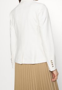 Una donna che indossa un blazer bianco aderente con bottoni dorati sui polsini e una gonna beige plissettata, vista da dietro.