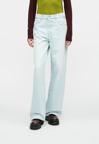 Ανοιχτό μπλε τζιν παντελόνι με φαρδιά μπατζάκια και ψηλή μέση, με απαλή υφή ντένιμ, συνδυασμένο με πράσινο και βαθυκόκκινο μακρυμάνικο τοπ και χοντρά μαύρα παπούτσια.