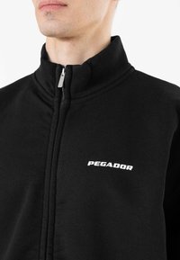 Crna jakna na zip, izrađena od mekanog materijala, ima visoki ovratnik, glatku teksturu i bijeli logo "PEGADOR" s lijeve strane.