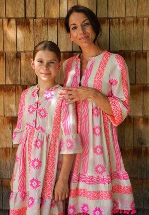 Frau und Mädchen stehen Seite an Seite vor einer Holzwand, tragen passende rosa-weiße gemusterte Kleider, Frau legt ihre Hand auf die Schulter des Mädchens.