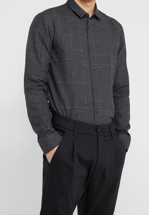 Mann trägt ein dunkelgraues Hemd mit dünnen hellen Gittern und schwarze plissierte Hosen, die Hände in den Taschen.