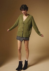 Groene gebreide cardigan met een V-hals, vijf bruine knopen aan de voorkant en een geribbelde zoom. Gecombineerd met een zwart-witte geruite rok en zwarte laarzen.