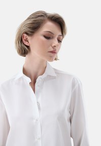Camicia bianca con colletto classico, caratterizzata da bottoni in perla e una texture del tessuto liscia, presentata in una vista ravvicinata.