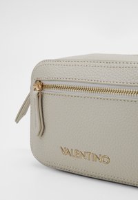 Krémová texturovaná kožená taštička se zlatým zipem, vybavená malým koženým táhlem a embosovaným logem "VALENTINO".