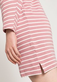 Vestido riscado rosa com riscas brancas horizontais, apresentando uma fenda lateral e material de algodão macio. Primeira plano do detalhe da manga e da bainha.