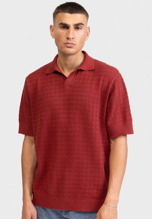 Νεαρός άνδρας που φοράει κόκκινο σκούρο πόλο με κοντά μανίκια και ανάγλυφη υφή, καθώς και ανοιχτό μπλε τζιν, στέκεται μπροστά σε απλό ανοιχτό φόντο.