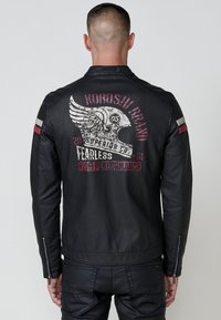 Uomo che indossa una giacca di pelle nera con un logo a elmo con ali e il testo "Koroshi Brand, Superior Co, Fearless, High Performance" sulla schiena.