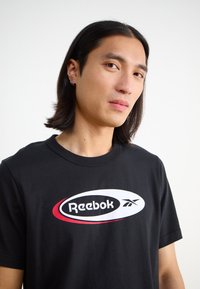 Fekete pamut póló rövid ujjal és kerek nyakkivágással, elöl fehér és piros Reebok logóval, ovális formában.