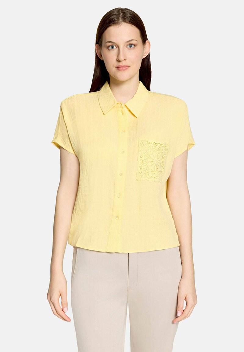 Femme portant une chemise boutonnée à manches courtes jaune pâle avec une poche décorative en dentelle, associée à un pantalon beige clair, debout devant un fond blanc.