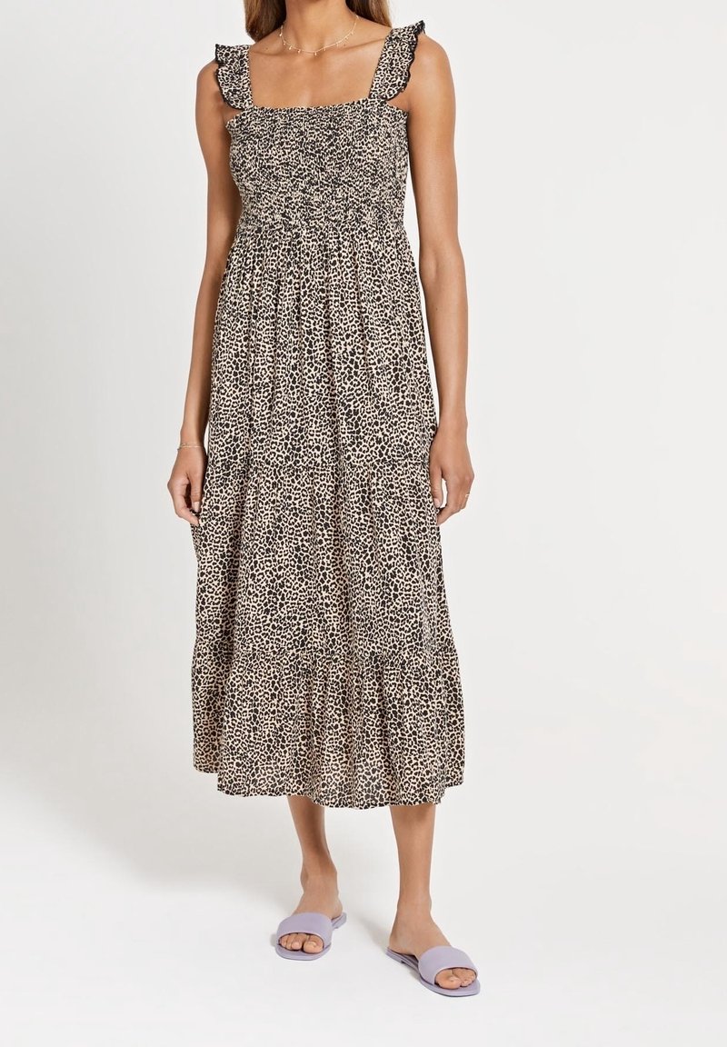 Femme portant une robe midi sans manches à imprimé léopard avec des bretelles à volants, associée à des sandales ouvertes lavande, sur un fond uni.