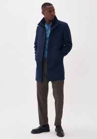 Abrigo de lana azul marino con cuello alzado, que presenta un diseño de un solo botón y tres botones. Combinado con pantalones a cuadros marrones y zapatos negros.