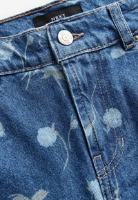 Tejido de mezclilla en azul claro, con un estampado floral. Incluye un cierre de botón plateado y costuras en color marrón claro a lo largo de las costuras. Etiqueta de la marca visible.