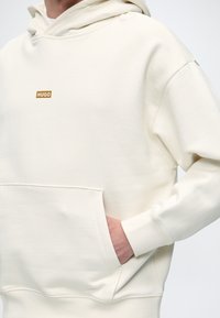 Personne portant un sweat à capuche couleur crème avec un logo "HUGO" sur la poitrine, main placée à l'intérieur de la poche kangourou avant.