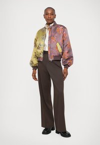 Chaqueta bomber en púrpura y oliva con patrones florales; textura acolchada, puños acanalados y cierre con cremallera. Combinada con pantalones marrones.