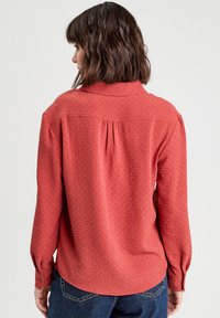 Blouse rouge texturée avec une fente au dos, des poignets boutonnés et un col, assortie à un pantalon en denim bleu pour un contraste.