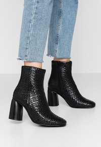Botas de tobillo negras y texturizadas con un patrón de piel de cocodrilo, punta afilada y tacón grueso. Llevadas con unos jeans de mezclilla azul cortos.
