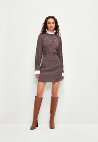 Robe mini à motifs marron avec manches longues et col en dentelle blanc, associée à des bottes marron jusqu'aux genoux et des talons noirs.