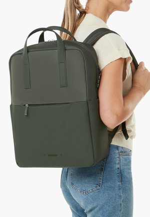 Frau trägt einen dunkelgrünen rechteckigen Rucksack mit zwei Griffen und einer vorderen Reißverschlusstasche, bekleidet mit einem beigen Oberteil und blauen Jeans.