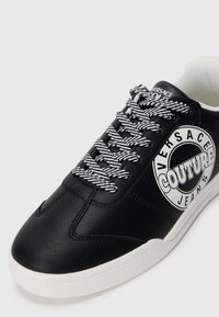 Črne nizke superge z belim podplatom, črtastimi črno-belimi vezalnimi trakovi in logotipom Versace Jeans Couture na strani in jeziku.