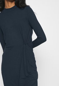 Mörk marinblå långärmad klänning med hög halsringning och vriden sidodetalj, tillverkad av ett stretchigt material med en mjuk textur.