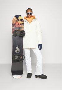Weiße Snowboardjacke mit orangefarbenem Innenfutter, kombiniert mit weißen Snowboardhosen. Das Snowboard hat ein farbenfrohes Design und schwarze Bindungen.