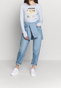Camicia a maniche lunghe a righe azzurro chiaro con logo dorato, abbinata a un maglione azzurro chiaro legato in vita e jeans in denim chiaro strappati. Sneakers bianche.
