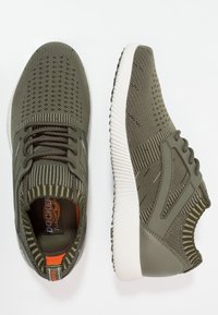 Scarpe sportive verde oliva con tomaia in rete testurizzata, colletto in maglia elastica e suola in gomma bianca. Presentano lacci e dettagli laterali.