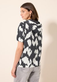 Camisa de manga corta con capucha que presenta un patrón abstracto en blanco y negro, tejido texturizado y un ajuste relajado. Bajo ligeramente suelto, jeans grises.