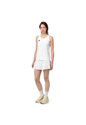 Giovane donna in piedi, indossa un top bianco senza maniche e una gonna abbinata, sneakers beige e calzini bianchi, su uno sfondo bianco uniforme.