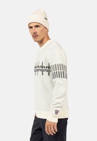 Weißer Strickpullover mit einem schwarz-grauen geometrischen Muster über der Brust. Kombiniert mit einer gerippten weißen Mütze für zusätzliche Wärme.