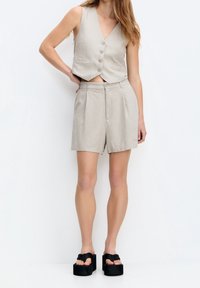 Veste courte beige avec boutons sur le devant, associée à un short beige taille haute. Portée avec des sandales à plateforme noires, sur un fond uni.