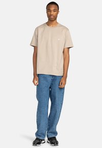 Beige Baumwoll-T-Shirt mit Rundhalsausschnitt, kurzen Ärmeln und einem kleinen Logo auf der Brust, kombiniert mit lockeren blauen Jeans und schwarzen Sneakers.