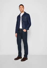 Muškarac stoji odjeven u tamnoplavu traper jaknu, bijelu majicu, tamne traperice i smeđe kožne cipele naspram jednostavne sive pozadine.