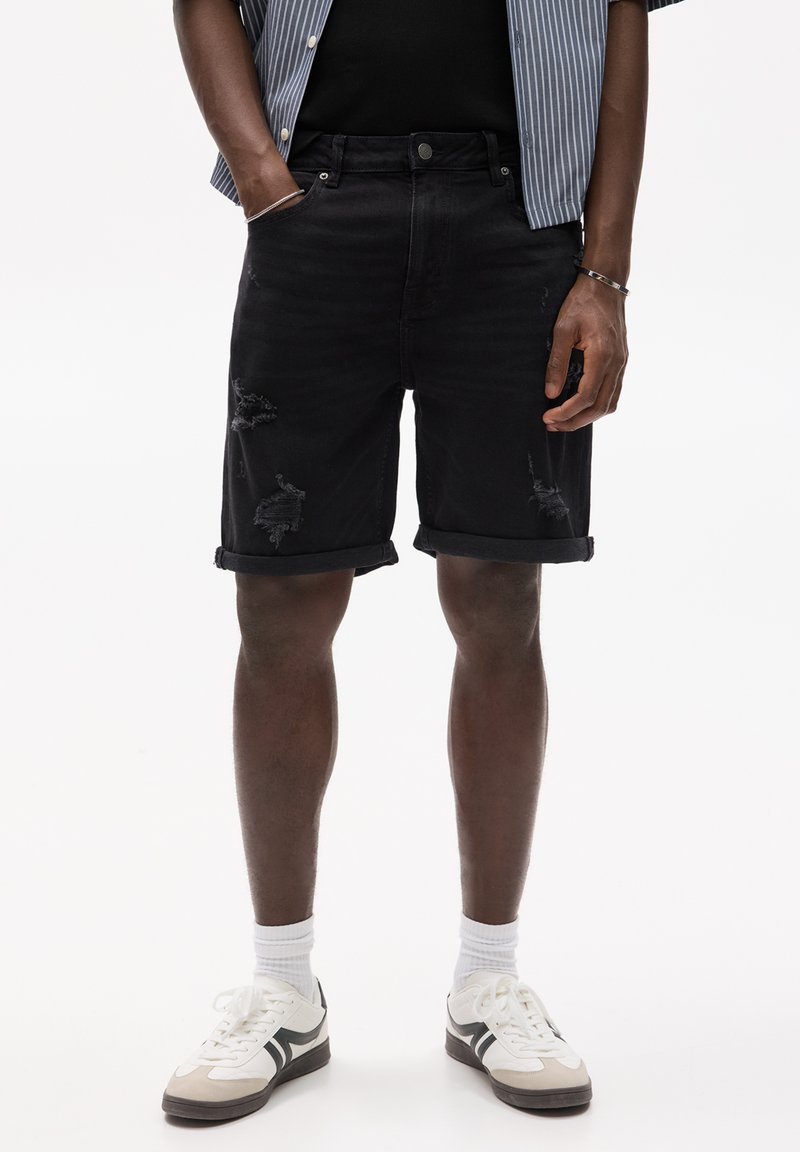 Shorts en denim noir avec des détails déchirés et des manches retroussées, portés avec des baskets blanches et des chaussettes gris clair. Le mannequin se tient dans un cadre neutre.