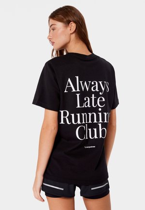 Kobieta o brązowych włosach, ubrana w czarne krótkie spodenki i czarną koszulkę z białym napisem „Always Late Running Club” na plecach, zwrócona w lewo.