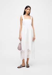 Γυναίκα που φοράει λευκό αμάνικο μεσαίο φόρεμα με δαντελωτό τελείωμα, κρατώντας μια μικρή ροζ τσάντα χειρός και φορώντας σανδάλια φλατ με μοτίβο φιδιού.