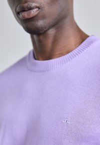 Ljuslila stickad tröja med rund halsringning och ribbad mudd; liten vit logotyp på bröstet; mjuk textur.