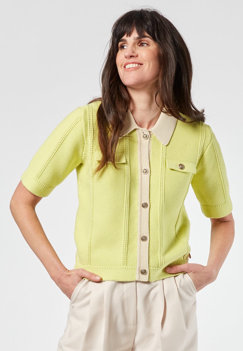 Kurze Strickjacke mit kurzen Ärmeln, hellgelb mit kontrastierendem beigen Kragen, zwei Fronttaschen und Knöpfen in Goldoptik, mit strukturierten Stichdetails.